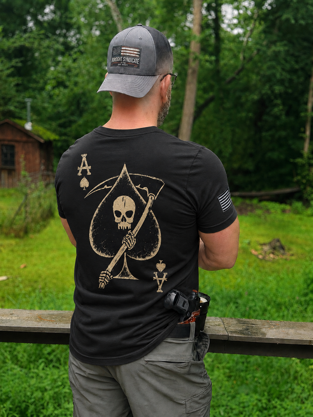 A man viewed from behind wearing a black t-shirt with a large beige Grim Reaper and Ace of Spades graphic on the back, standing outdoors in a grassy yard.
