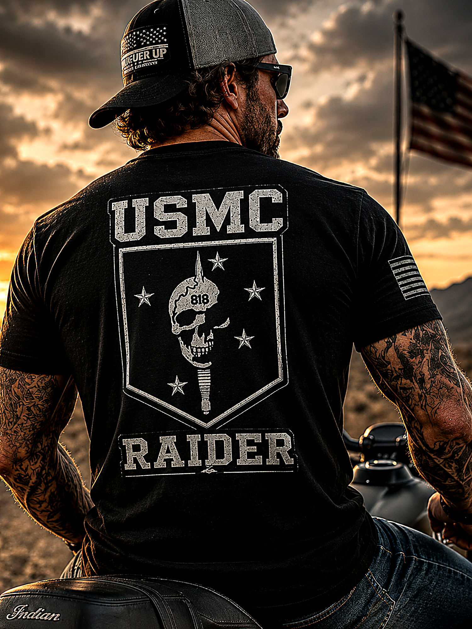 Man wearing a USMC Raider t-shirt with a skull design, sitting on a motorcycle against a sunset sky.